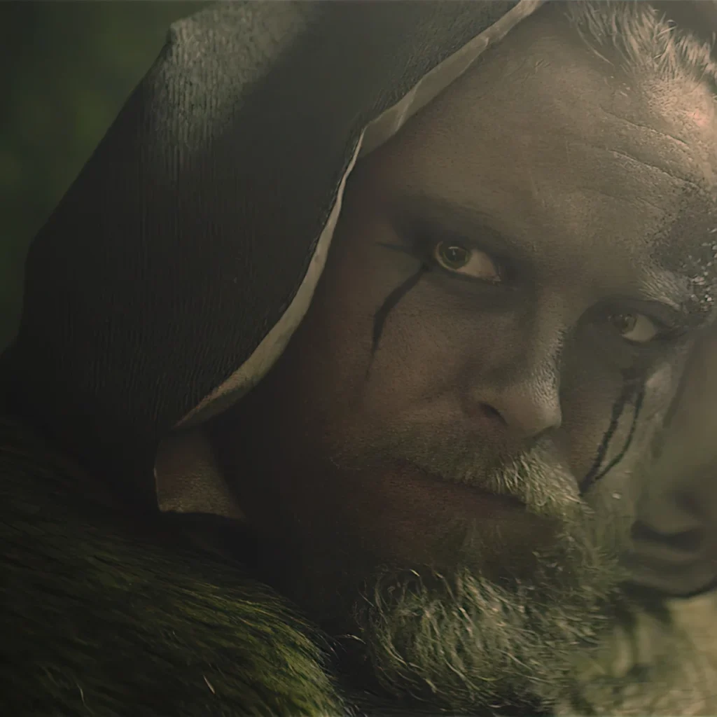 A man styled as a Viking, wearing a hood and dark makeup around his eyes, stares intensely at the camera; the dark background and Nordic styling create an atmosphere of mystery and dread.
