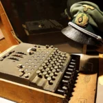 Fotografia muzealnej ekspozycji: w drewnianej skrzyni stoi historyczna maszyna szyfrująca Enigma z obudową z klawiaturą literową i pokrętłami, obok spoczywa zielona czapka oficerska z insigniami armii niemieckiej.