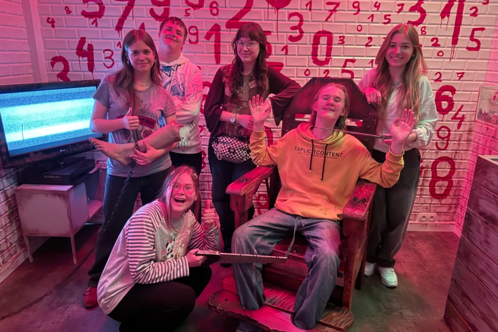 A group of teenagers in a horror-themed escape room inspired by “Saw”; one person sits in a torture chair with fake handcuffs and a saw, while the rest smile after successfully solving the puzzle. In the background, a wall marked with bloody numbers.