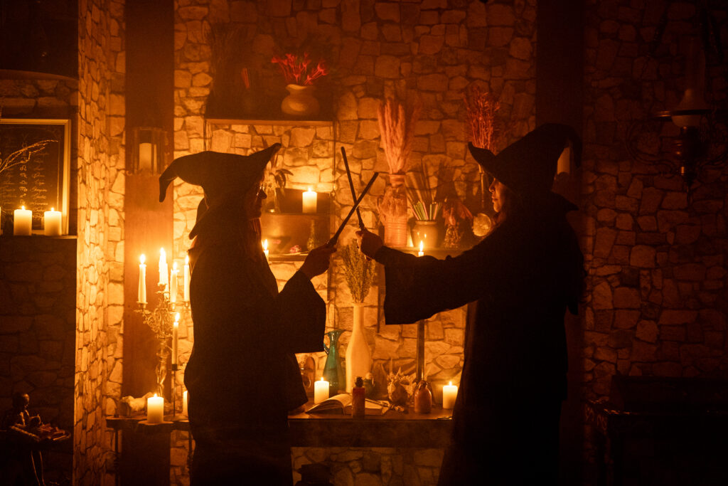 Dwie osoby w strojach czarownic stoją naprzeciwko siebie w przyciemnionym, magicznie oświetlonym pomieszczeniu. Trzymają różdżki skrzyżowane w powietrzu, jakby brały udział w pojedynku czarodziejów. W tle widoczne są kamienne ściany, zapalone świece i półki z magicznymi rekwizytami.