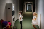 Dwie dziewczynki biorą udział w zabawie z fotobudką, jedna pozuje przed obiektywem, druga skacze z radości; obok leżą kolorowe przebrania.