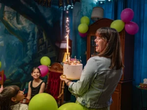 Kobieta wnosi tort urodzinowy z zimnym ogniem do udekorowanej sali, a dzieci uśmiechają się i czekają na rozpoczęcie świętowania.