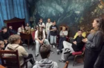 Grupa uczniów w sali stylizowanej na magiczną bibliotekę uważnie słucha kobiety prowadzącej zajęcia, niektórzy siedzą, inni stoją, atmosfera skupienia i zaciekawienia.