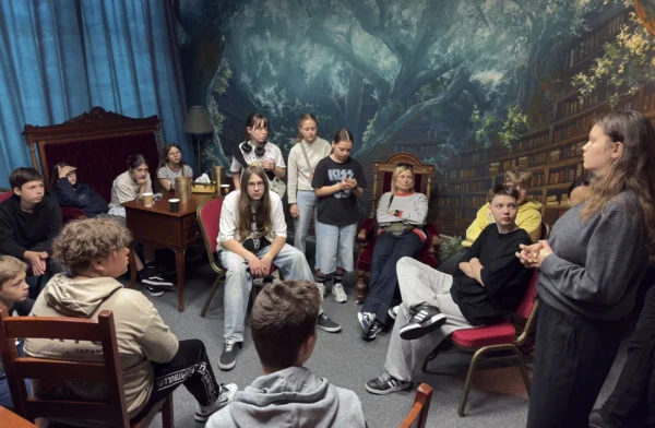 Grupa uczniów w sali stylizowanej na magiczną bibliotekę uważnie słucha kobiety prowadzącej zajęcia, niektórzy siedzą, inni stoją, atmosfera skupienia i zaciekawienia.