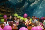 Kilkoro dzieci siedzi przy stole podczas urodzin w sali z balonami, kolorowymi przekąskami i owocami; w tle magiczna biblioteka z malowanymi drzewami.