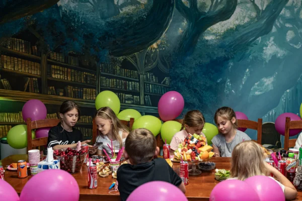 Kilkoro dzieci siedzi przy stole podczas urodzin w sali z balonami, kolorowymi przekąskami i owocami; w tle magiczna biblioteka z malowanymi drzewami.