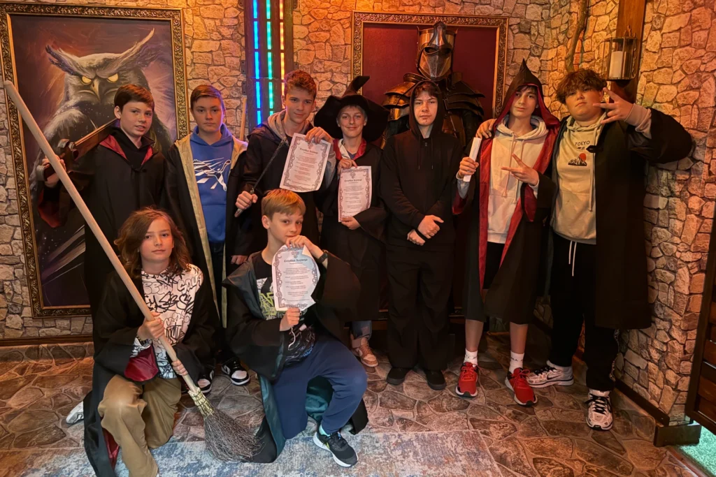 Grupa chłopców w strojach czarodziejów po udanej grze w pokoju zagadek o tematyce magicznej, trzymają certyfikaty i różdżki, w tle obraz sowy i zbroja rycerska.
