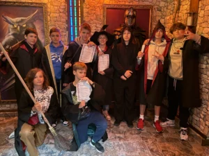Grupa chłopców w strojach czarodziejów po udanej grze w pokoju zagadek o tematyce magicznej, trzymają certyfikaty i różdżki, w tle obraz sowy i zbroja rycerska.