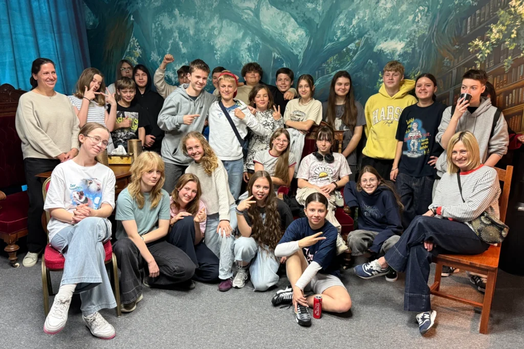Duża grupa uczniów wraz z nauczycielkami po zakończonej grze w pokoju zagadek, wszyscy uśmiechnięci i zrelaksowani, w tle malowidło przedstawiające magiczny las.