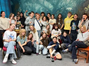 Duża grupa uczniów wraz z nauczycielkami po zakończonej grze w pokoju zagadek, wszyscy uśmiechnięci i zrelaksowani, w tle malowidło przedstawiające magiczny las.