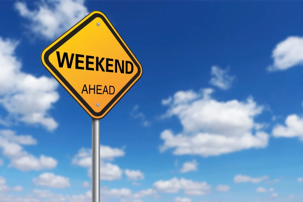 Żółty znak drogowy z napisem "WEEKEND AHEAD" na tle błękitnego nieba z białymi chmurami - symbol nadchodzącego długiego weekendu w Warszawie