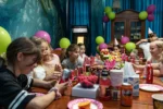 Grupa dzieci świętuje urodziny przy udekorowanym stole w escape roomie. Na stole stoją napoje, owoce i przekąski, a w tle widać kolorowe balony i dekoracje. Dzieci uśmiechają się, rozmawiają i cieszą wspólną zabawą.