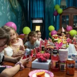 Grupa dzieci świętuje urodziny przy udekorowanym stole w escape roomie. Na stole stoją napoje, owoce i przekąski, a w tle widać kolorowe balony i dekoracje. Dzieci uśmiechają się, rozmawiają i cieszą wspólną zabawą.