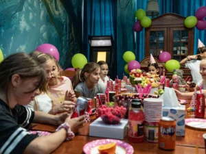 Grupa dzieci świętuje urodziny przy udekorowanym stole w escape roomie. Na stole stoją napoje, owoce i przekąski, a w tle widać kolorowe balony i dekoracje. Dzieci uśmiechają się, rozmawiają i cieszą wspólną zabawą.