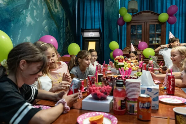 Grupa dzieci świętuje urodziny przy udekorowanym stole w escape roomie. Na stole stoją napoje, owoce i przekąski, a w tle widać kolorowe balony i dekoracje. Dzieci uśmiechają się, rozmawiają i cieszą wspólną zabawą.